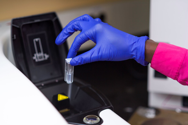 A safety-gloved hand pinching a small vial of liquid above a centrifuge