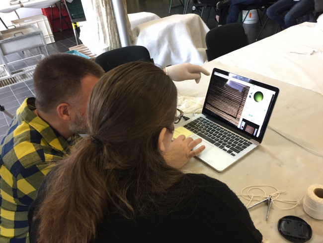 Two people sit at a desk and look at a laptop screen that shows the wooden tables and the RTI interface that lets them move light around on the image of the tablet.