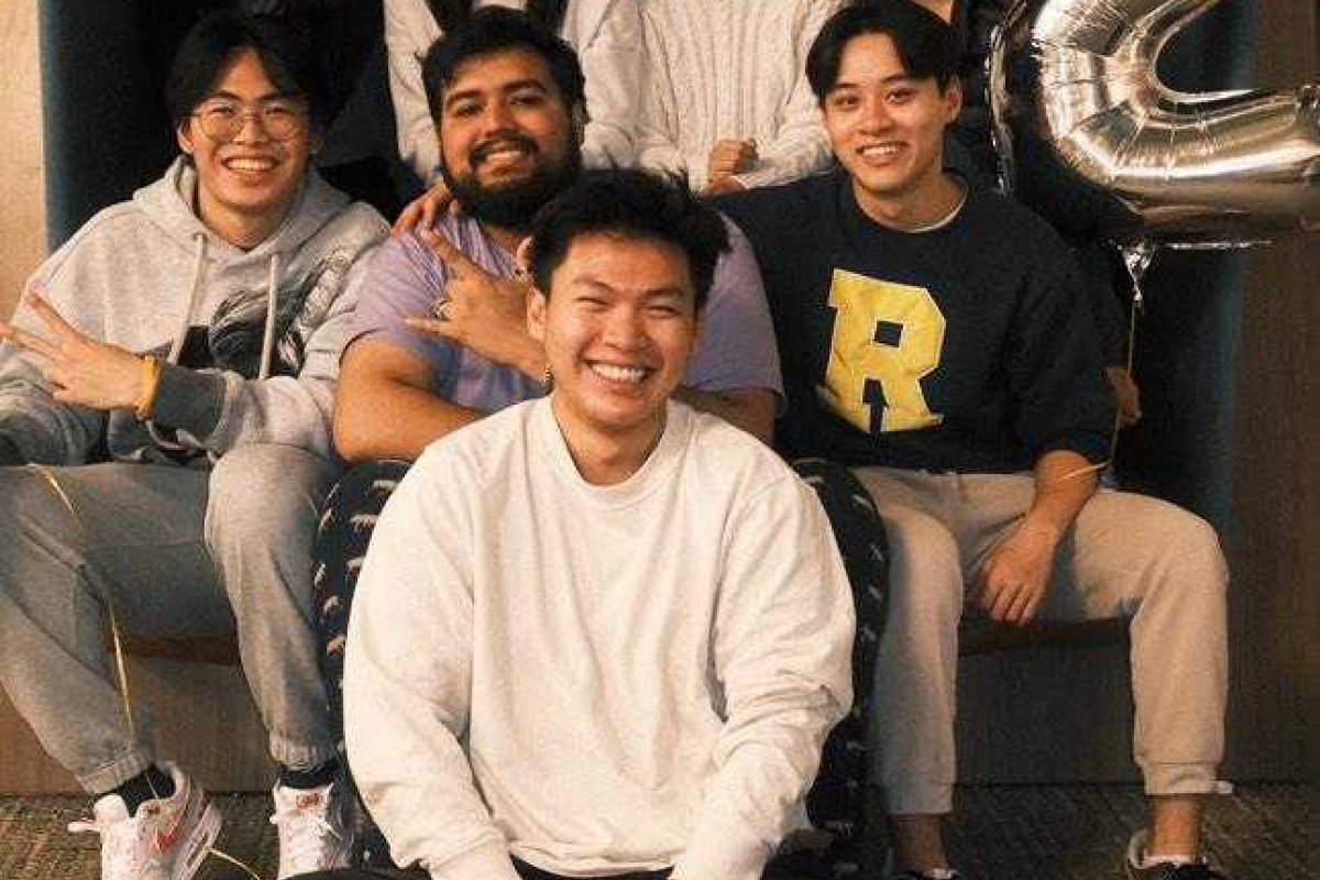Group photo of eight smiling individuals sitting together indoors. Four people are seated in the back row, and four are in the front, with one person sitting cross-legged on the floor. A large silver balloon in the shape of the number '2' is visible on the right side of the image. The group appears to be enjoying a relaxed and cheerful moment.