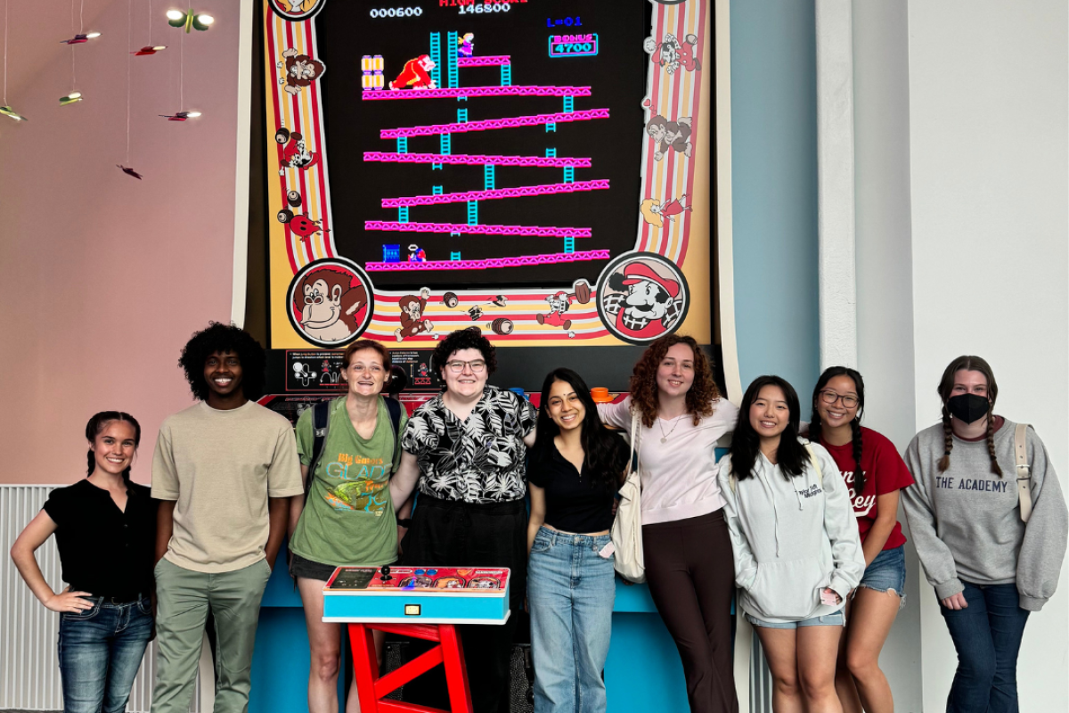 A group of nine people stand in front of a large Donkey Kong arcade machine. Some are smiling and some are casually posing. They're indoors with a gray floor and white walls.