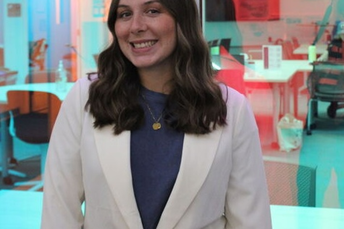 Liv Riz wearing a white blazer standing in front of a blue, semi-transparent screen 