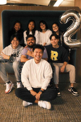 Group photo of eight smiling individuals sitting together indoors. Four people are seated in the back row, and four are in the front, with one person sitting cross-legged on the floor. A large silver balloon in the shape of the number '2' is visible on the right side of the image. The group appears to be enjoying a relaxed and cheerful moment.