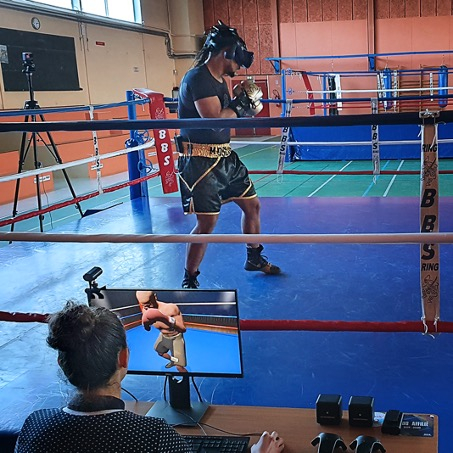 A person wearing a VR headset and boxing gloves trains in a boxing ring while someone monitors their progress on a screen nearby.