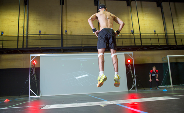 A shirtless athlete wearing motion-tracking sensors jumps in front of a large screen displaying his movements, while a coach observes from the side in an indoor sports facility.