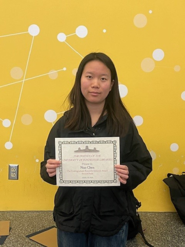 Student, Nuo Chen standing against a yellow wall holding an award certificate