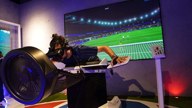 A person wearing a VR headset leans forward on a flight simulator setup with a fan blowing air, in front of a large screen showing a virtual soccer stadium.