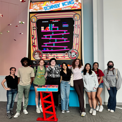 A group of nine people stand in front of a large Donkey Kong arcade machine. Some are smiling and some are casually posing. They're indoors with a gray floor and white walls.