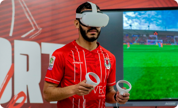 A man in a red soccer jersey is using a VR headset and controllers in front of a screen of a VR soccer field.