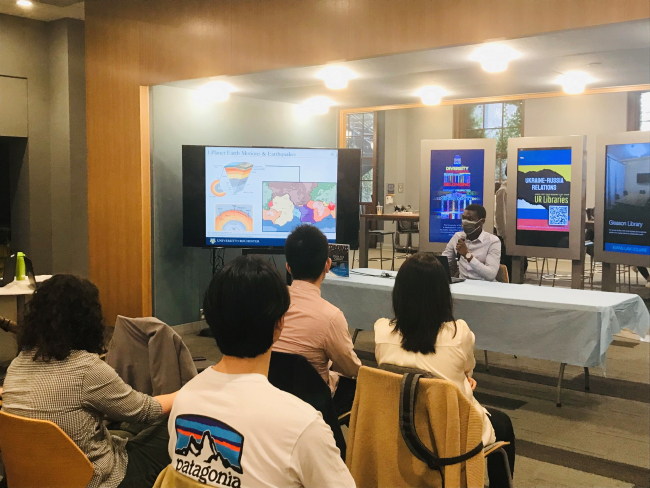 Student Jean-Joel Legre sitting at a table in Evans Lam Square talking about his project to presentation attendees