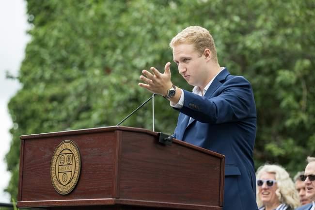 Consultant Elijiah Bader-Gergory standing next to a podium with his hand outstreched to the audience. 