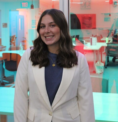 Liv Riz wearing a white blazer standing in front of a blue, semi-transparent screen 