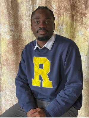 A person, Kwame Edu-Ansah, sits in front of draped fabric wearing a University of Rochester sweatshirt